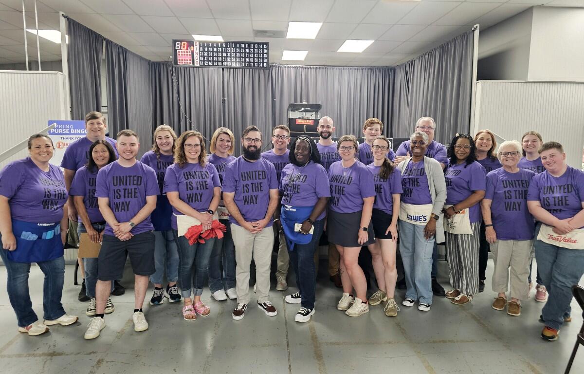 Group of about 25 people wearing matching purple shirts reading 'UNITED IS THE WAY' posing for a team photo in an indoor room.