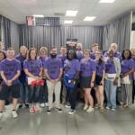 Group of about 25 people wearing matching purple shirts reading 'UNITED IS THE WAY' posing for a team photo in an indoor room.