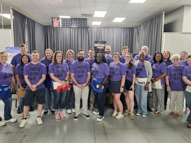 Group of about 25 people wearing matching purple shirts reading 'UNITED IS THE WAY' posing for a team photo in an indoor room.