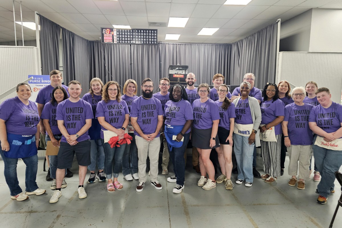 Group of about 25 people wearing matching purple shirts reading 'UNITED IS THE WAY' posing for a team photo in an indoor room.