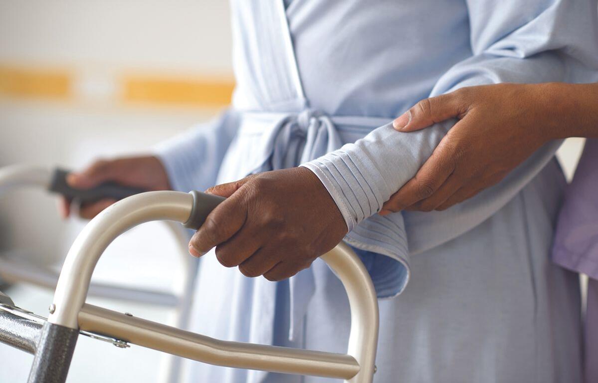 A patient in a light blue gown grips a walker handle while a caregiver steadies their arm for support.