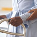 A patient in a light blue gown grips a walker handle while a caregiver steadies their arm for support.