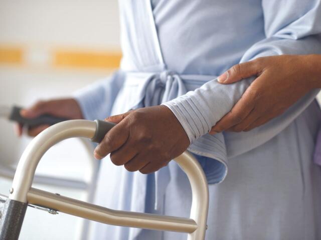 A patient in a light blue gown grips a walker handle while a caregiver steadies their arm for support.