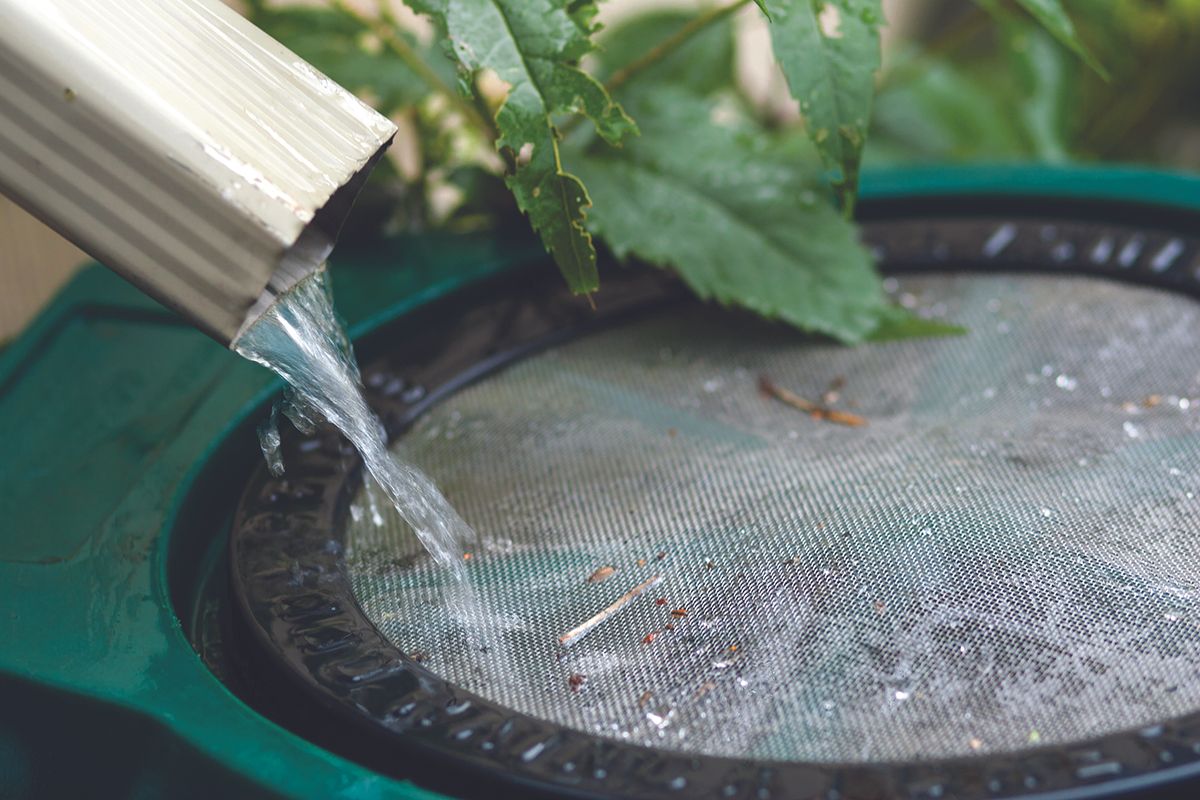 Water flows from a metal spout into a circular green mesh filter, with leaves resting on top and debris around the rim.