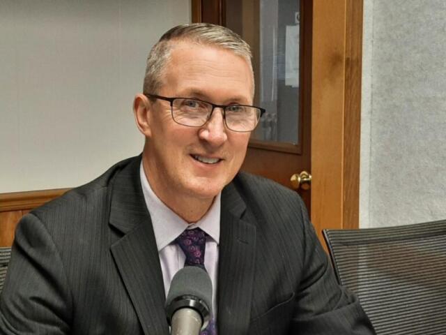 Smiling man in a dark suit and glasses speaks into a microphone in a meeting room.