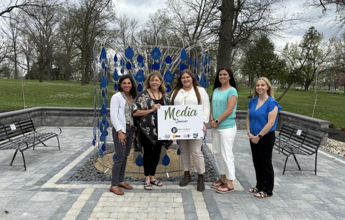 Five women pose in a park beside a blue teardrop sculpture and a 'Media Sponsor' sign at an outdoor event.