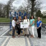 Five women pose in a park beside a blue teardrop sculpture and a 'Media Sponsor' sign at an outdoor event.