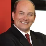 Smiling man in a dark suit and red tie, posing for a professional headshot with a red backdrop behind him.