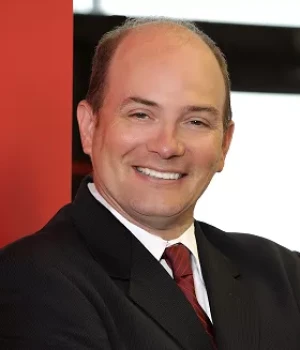 Smiling man in a dark suit and red tie, posing for a professional headshot with a red backdrop behind him.