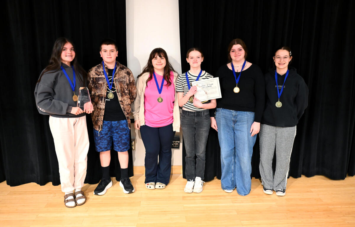 Six smiling teens stand on a stage with a black curtain backdrop, wearing medals; one holds a trophy and another a certificate.