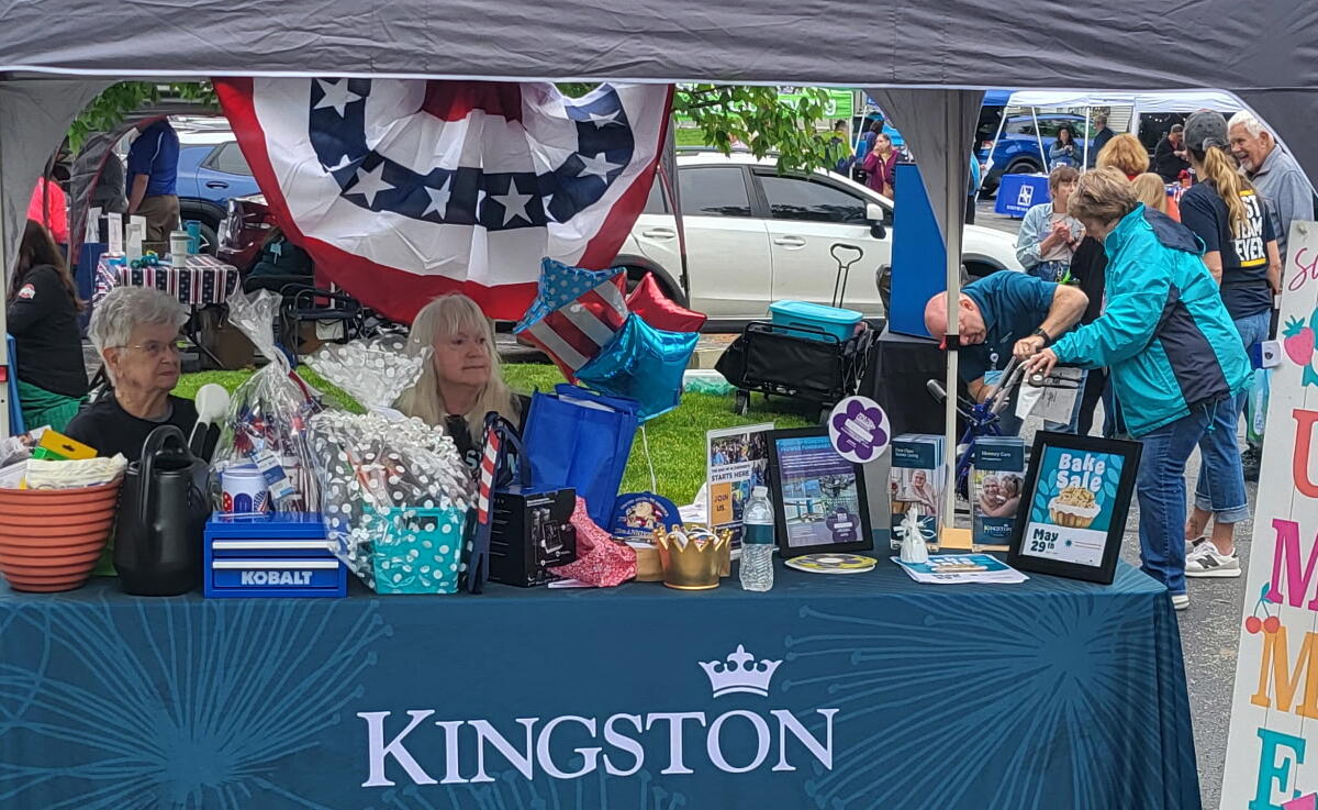 Outdoor Kingston booth at a community fair with tables, balloons, and gift baskets under a canopy, people browsing items.