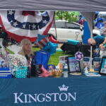 Outdoor Kingston booth at a community fair with tables, balloons, and gift baskets under a canopy, people browsing items.