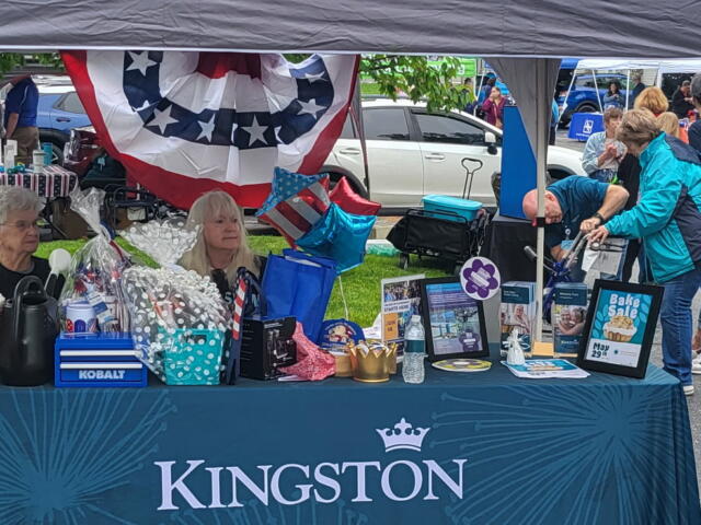 Outdoor Kingston booth at a community fair with tables, balloons, and gift baskets under a canopy, people browsing items.