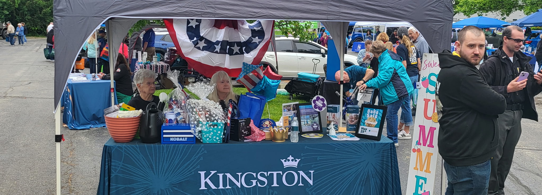 Outdoor Kingston booth at a community fair with tables, balloons, and gift baskets under a canopy, people browsing items.