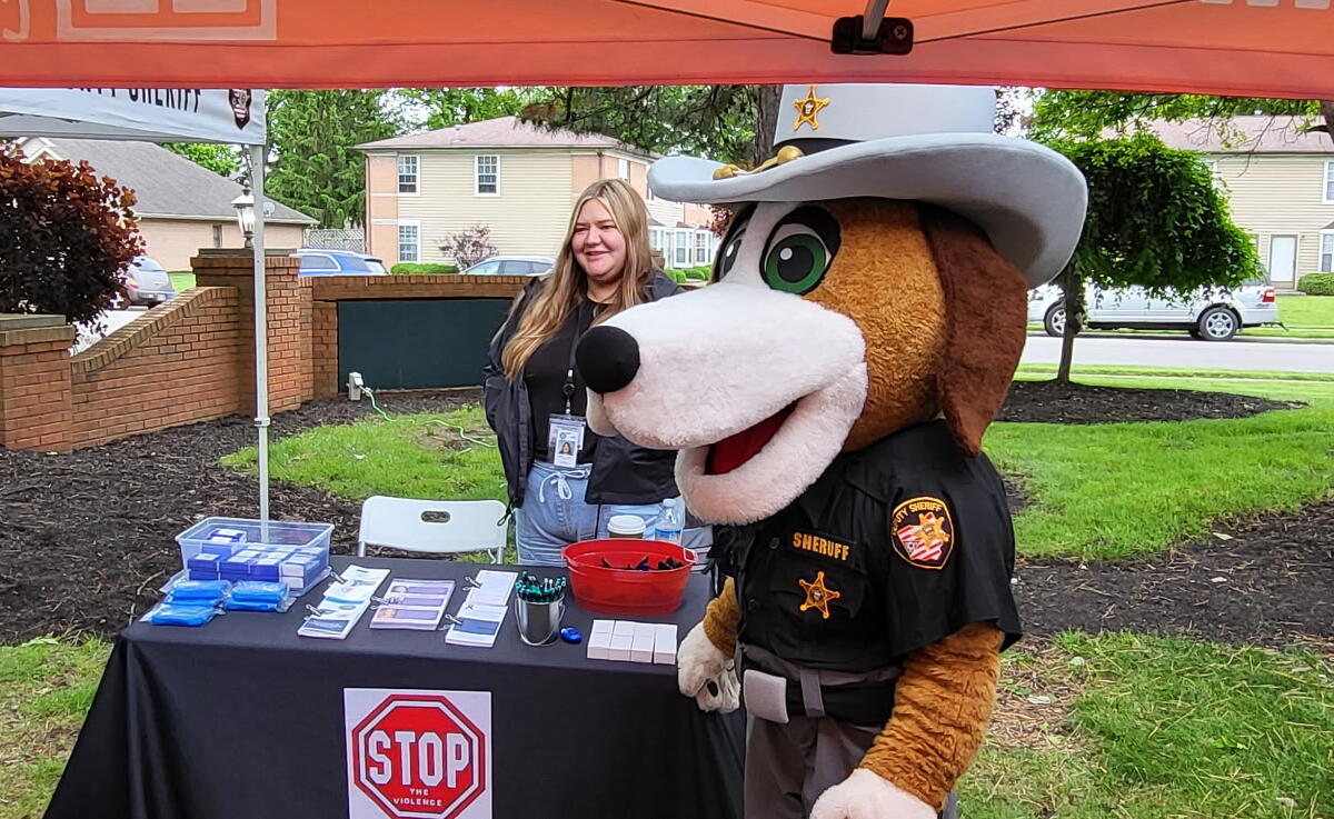 Costumed sheriff dog mascot at an outdoor information booth with a Stop the Violence sign on the table, beside a smiling woman volunteer.