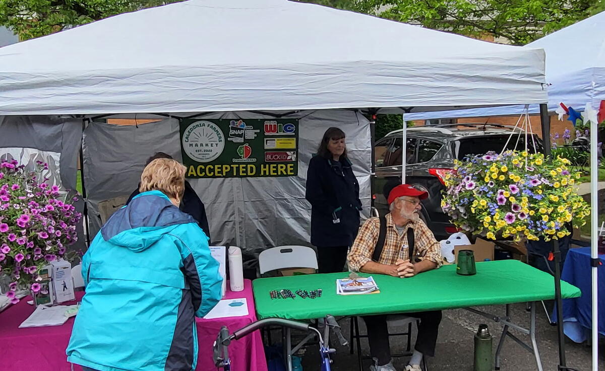 Outdoor community fair booth under white canopies with volunteers, a Heartland Hospice table, and bright flower baskets in bloom.