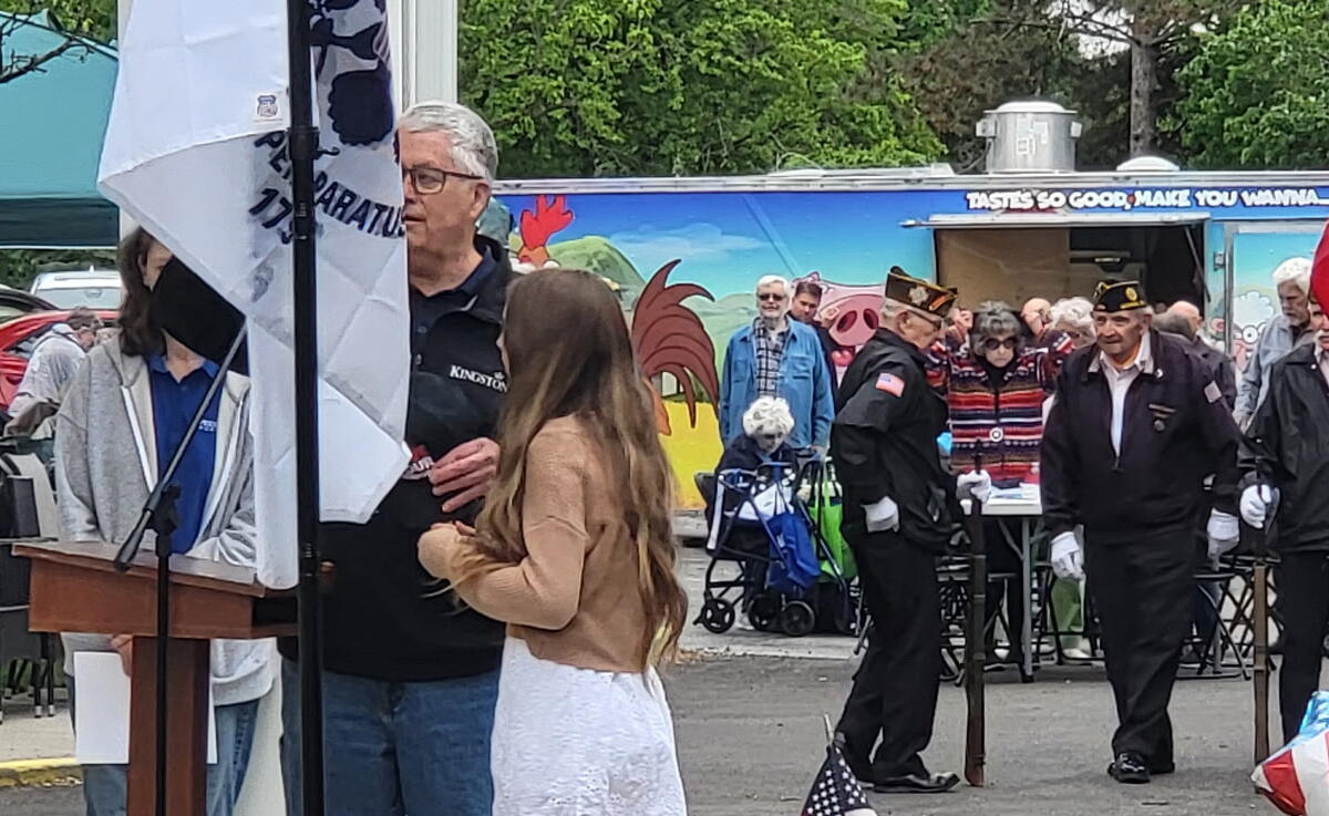 Outdoor ceremony with a speaker at a wooden podium, a young woman beside him, and a crowd of onlookers including veterans and flags in the scene.