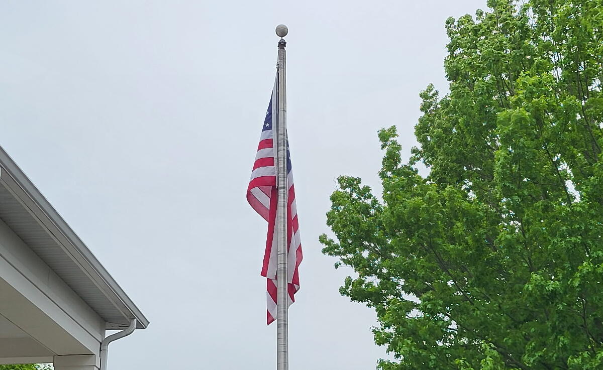 American flag on a tall flagpole beside a white building and lush green trees under a cloudy sky