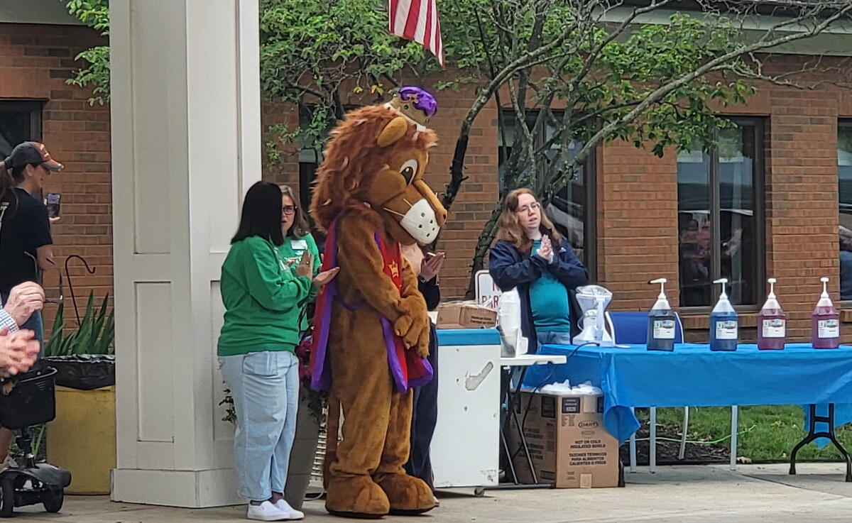 Outdoor community event; a person in a brown lion mascot stands by a table of hand sanitizer bottles while spectators watch.