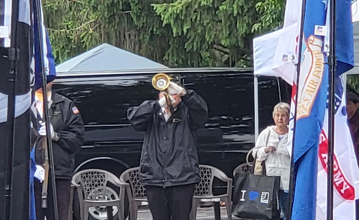 Person in dark jacket raises a brass megaphone at an outdoor ceremony with flags and a black van in the background.