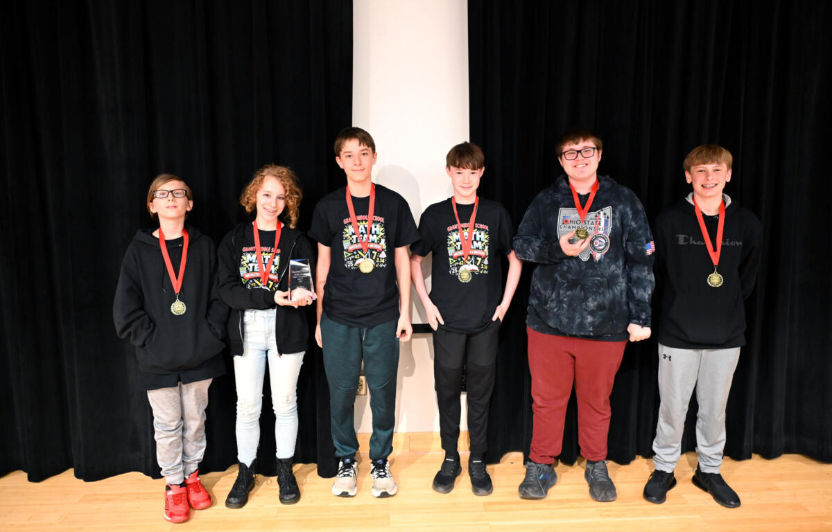 Six smiling students wearing medals stand on a stage with a black curtain behind them; one holds a trophy and plaque cups in the center-left.