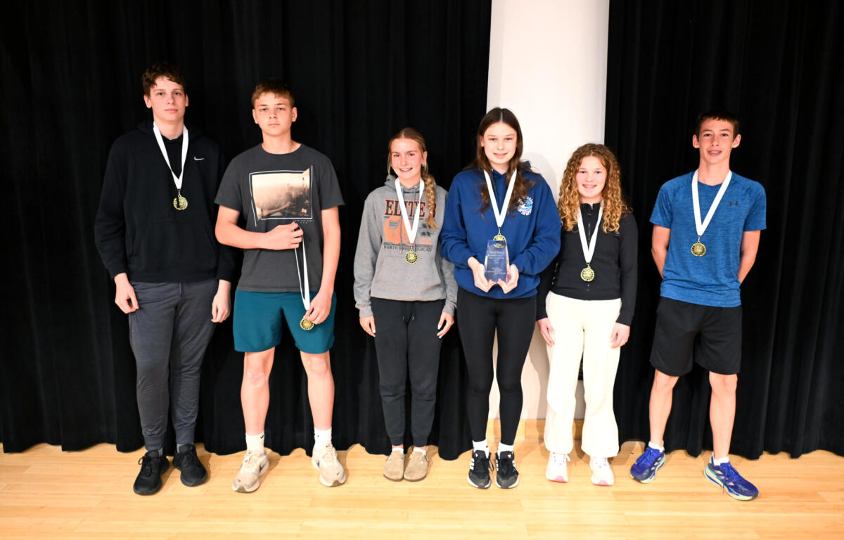 Group of seven young students on a stage wearing medals; one holds a trophy, posed in front of a black curtain.