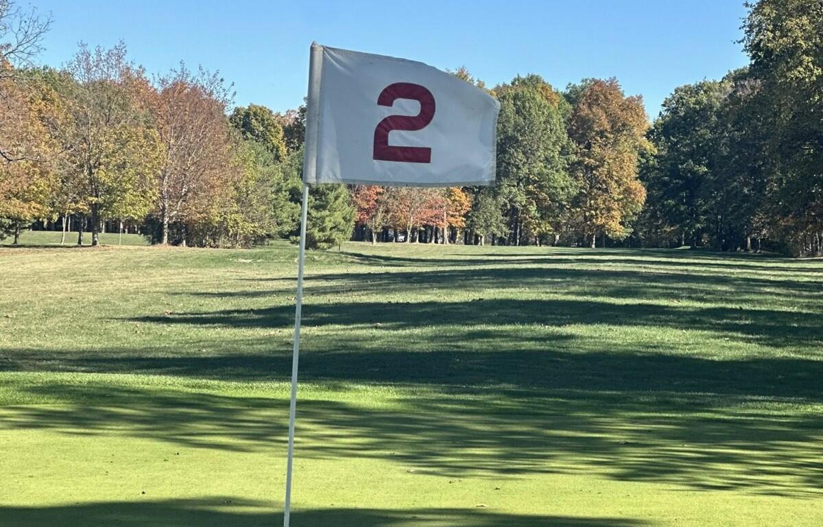 White flag with a red curved symbol on a golf course green, flagstick in the foreground against a sunny autumn backdrop.