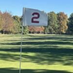 White flag with a red curved symbol on a golf course green, flagstick in the foreground against a sunny autumn backdrop.