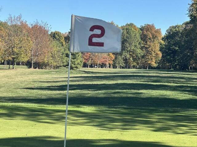 White flag with a red curved symbol on a golf course green, flagstick in the foreground against a sunny autumn backdrop.