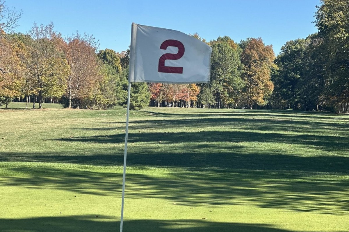 White flag with a red curved symbol on a golf course green, flagstick in the foreground against a sunny autumn backdrop.