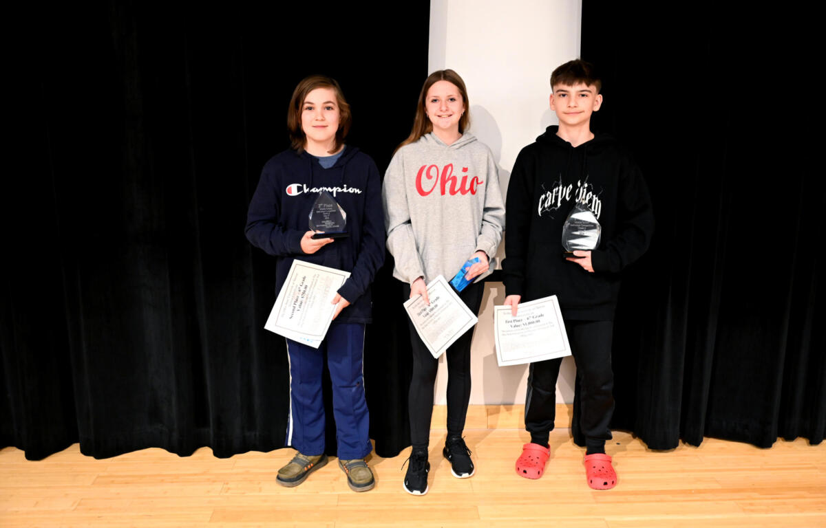 Three children stand on a stage with awards: left boy in navy Champion hoodie holding a trophy and certificate, middle girl in gray Ohio hoodie with papers, right boy in black hoodie with a trophy and certificates in front of a dark curtain.
