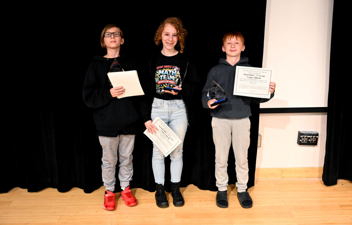 Three children stand on a stage holding awards and certificates, smiling; black curtain behind them and a wood floor visible.