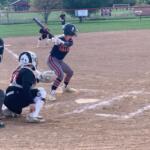Young batter in navy and orange swings at a pitch as the catcher and umpire crouch behind home plate in a youth softball game.