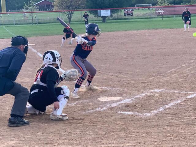Young batter in navy and orange swings at a pitch as the catcher and umpire crouch behind home plate in a youth softball game.