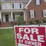 Red and white 'For Sale By Owner' sign in front yard of a brick house.