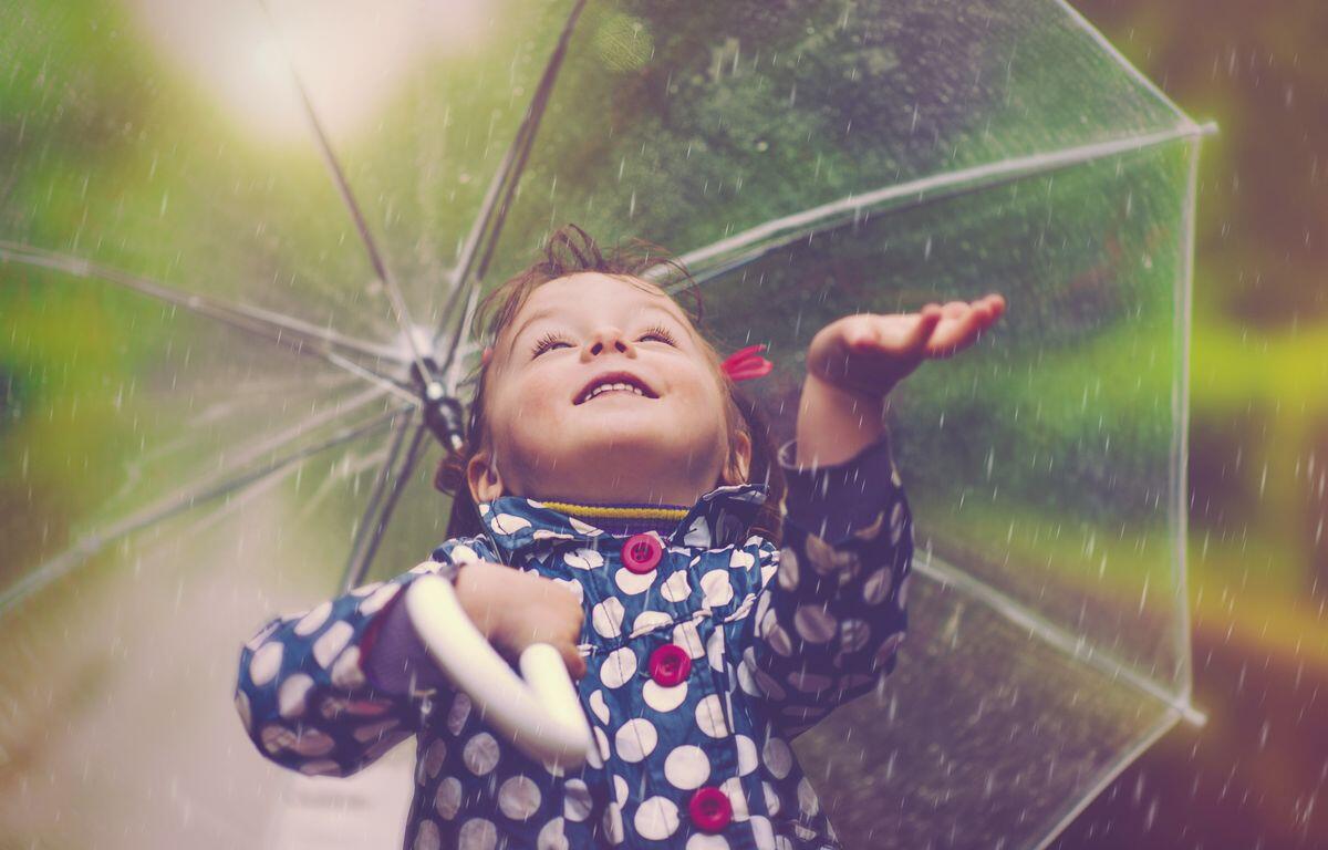 A smiling child raising an arm under a transparent umbrella in the rain, wearing a blue polka-dot raincoat.