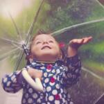 A smiling child raising an arm under a transparent umbrella in the rain, wearing a blue polka-dot raincoat.