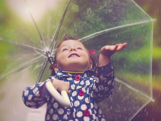 A smiling child raising an arm under a transparent umbrella in the rain, wearing a blue polka-dot raincoat.