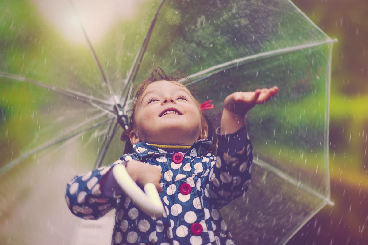 A smiling child raising an arm under a transparent umbrella in the rain, wearing a blue polka-dot raincoat.