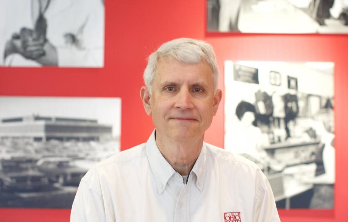 Portrait of a gray-haired man wearing a white button-down shirt, standing in front of a red wall with black-and-white photos behind him.