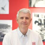 Portrait of a gray-haired man wearing a white button-down shirt, standing in front of a red wall with black-and-white photos behind him.