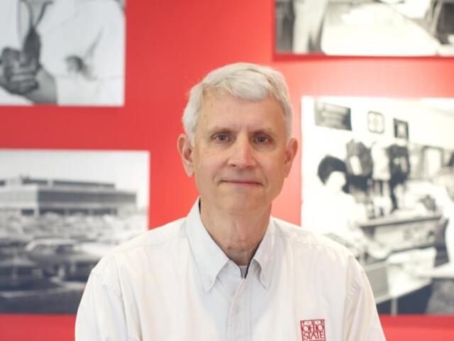 Portrait of a gray-haired man wearing a white button-down shirt, standing in front of a red wall with black-and-white photos behind him.