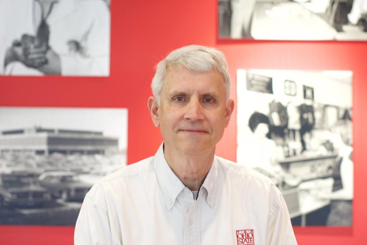 Portrait of a gray-haired man wearing a white button-down shirt, standing in front of a red wall with black-and-white photos behind him.
