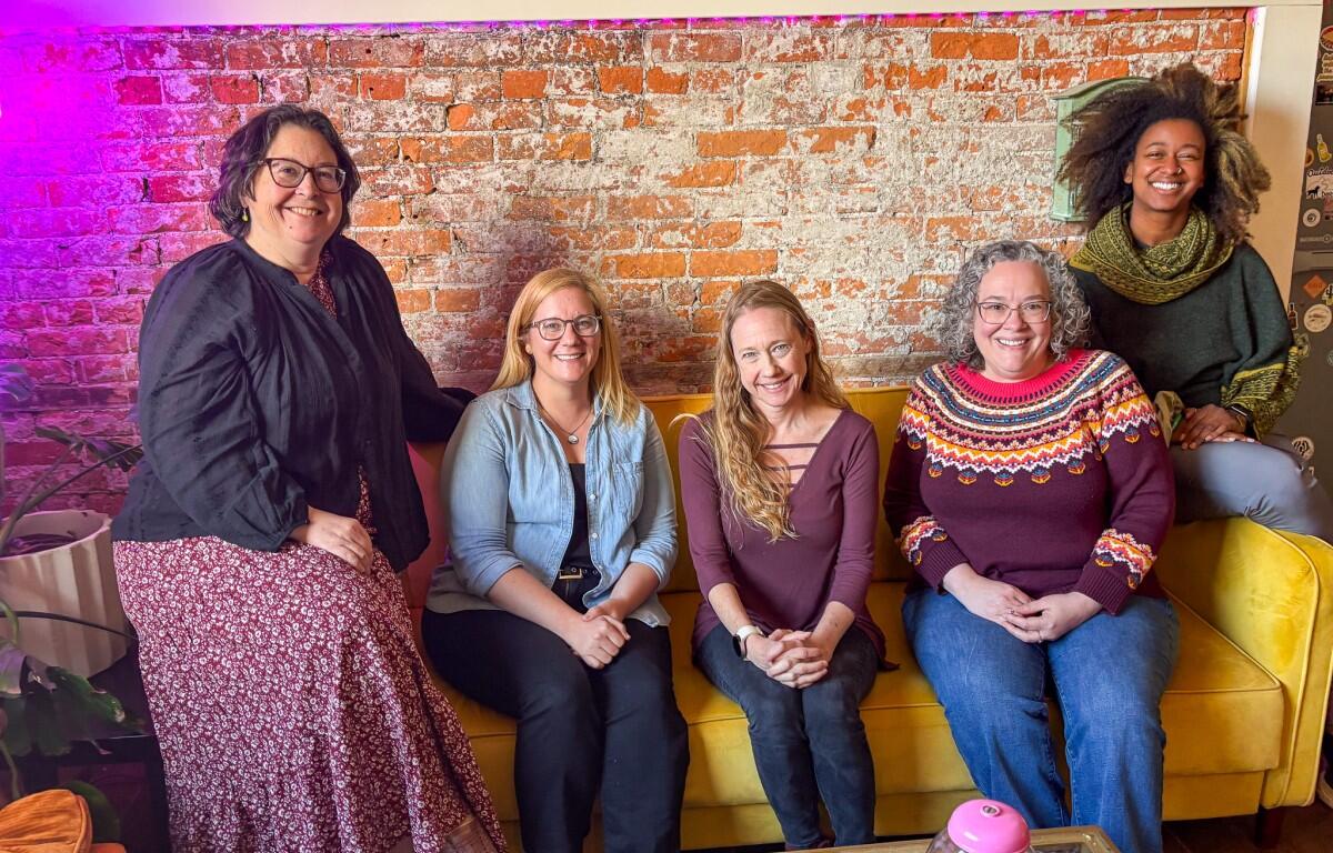 Five women smiling and seated on a mustard-yellow couch against a brick wall in a casual lounge setting.