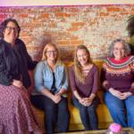 Five women smiling and seated on a mustard-yellow couch against a brick wall in a casual lounge setting.