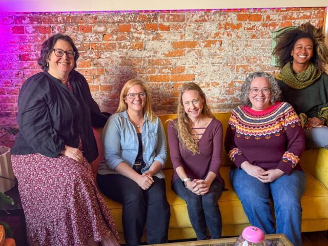 Five women smiling and seated on a mustard-yellow couch against a brick wall in a casual lounge setting.