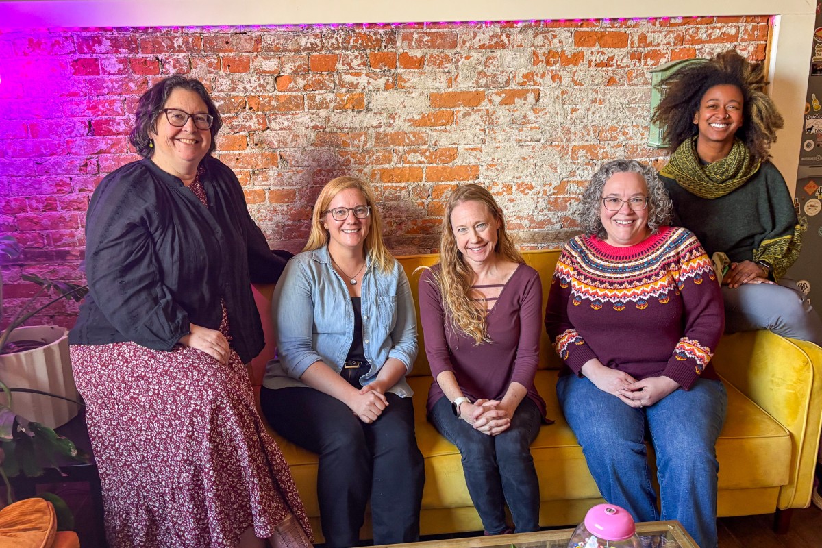Five women smiling and seated on a mustard-yellow couch against a brick wall in a casual lounge setting.