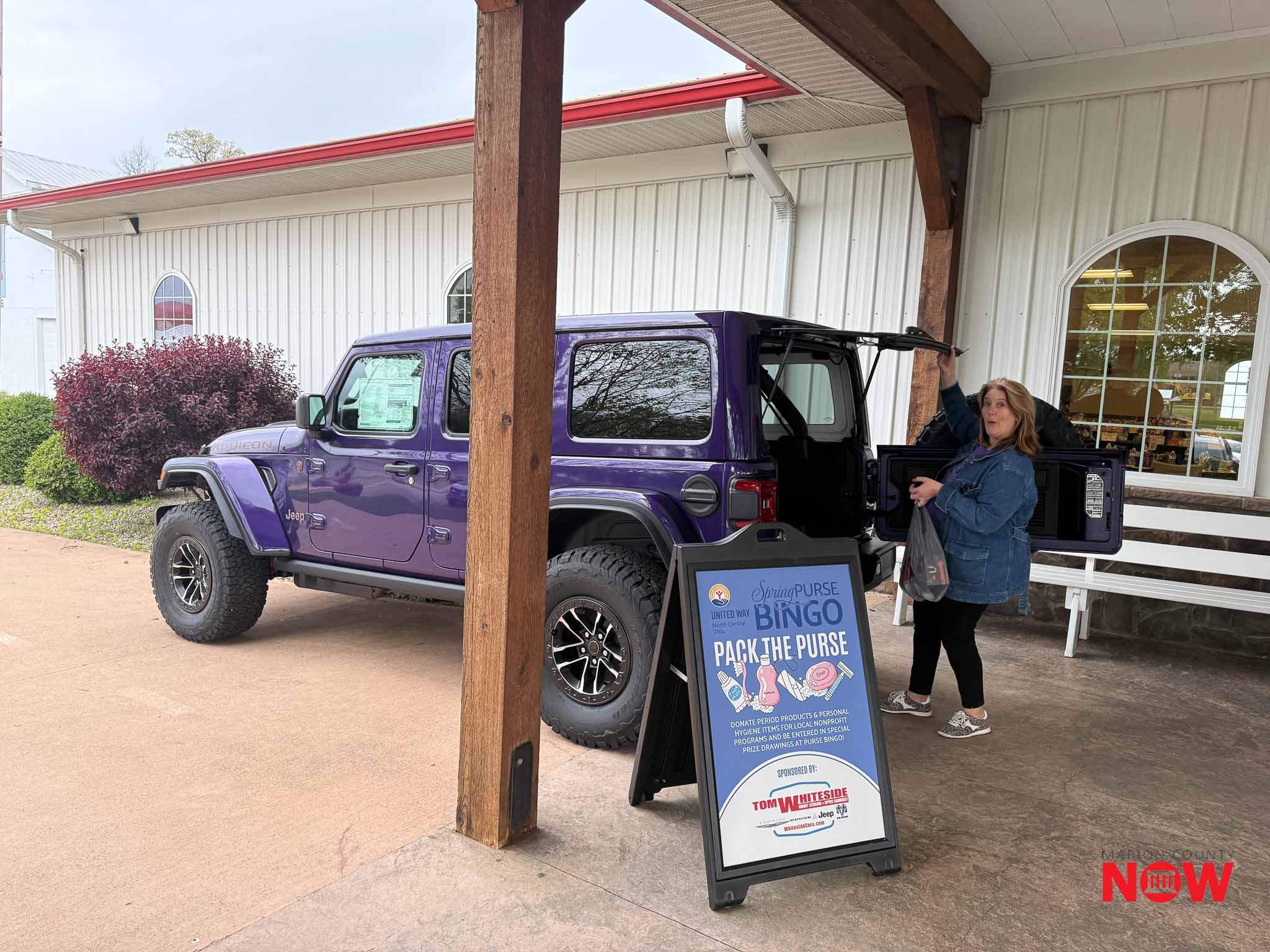 Purple Jeep with open tailgate parked under a wooden awning; a woman stands nearby placing items into the back, next to a promotional sign for a purse drive event.