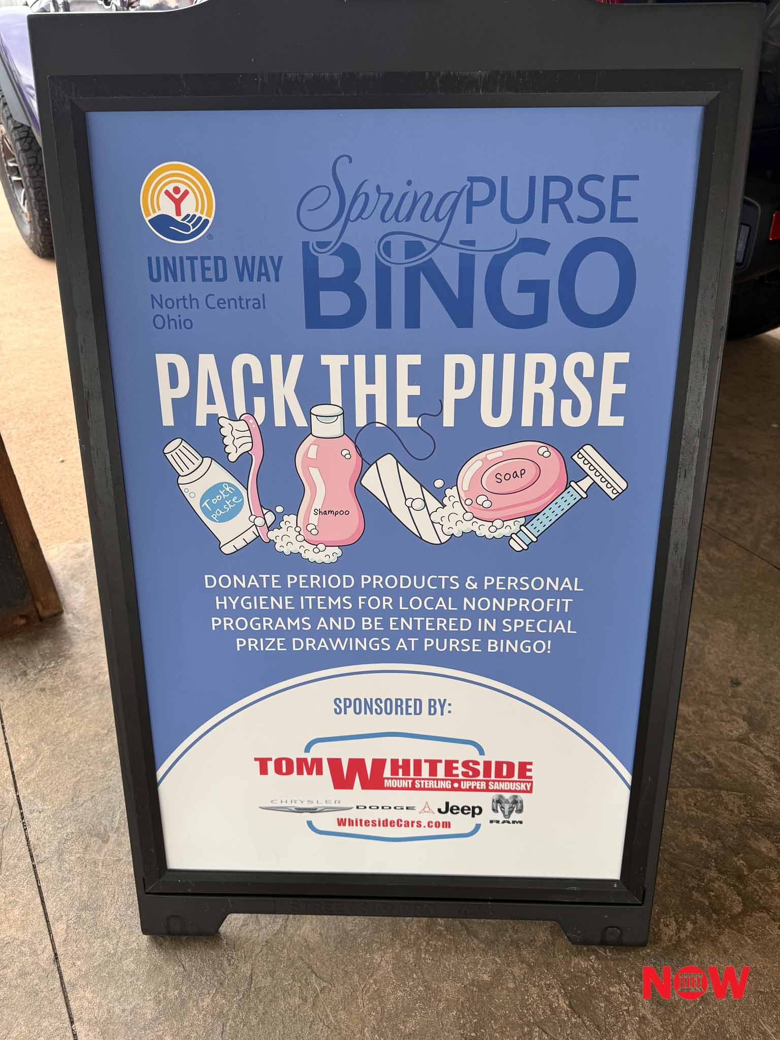 Blue promotional sign in a frame for Spring Purse Bingo by United Way, encouraging donations of hygiene products and period items (NOW sponsor logo visible).
