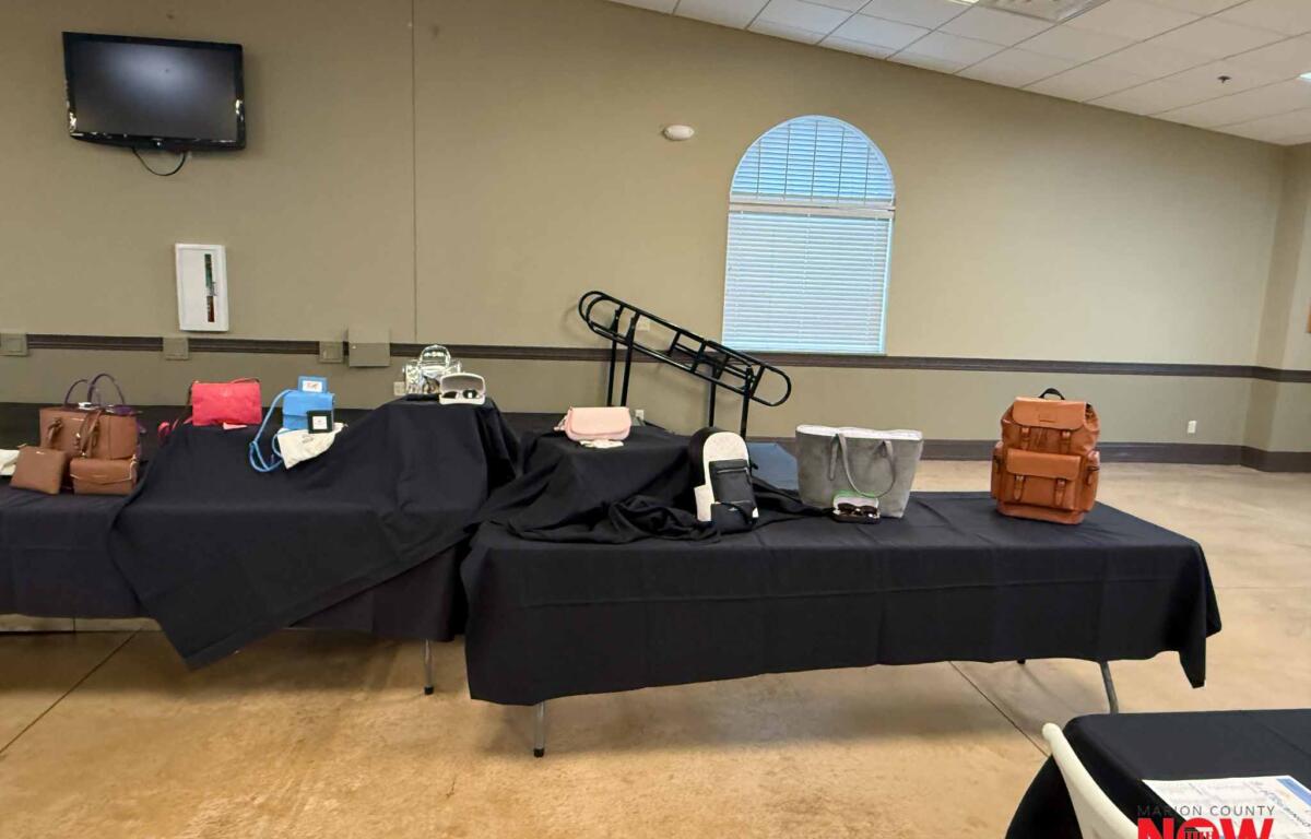 Event room with black-covered tables displaying bags and purses; wall-mounted TV and arched window in the background.